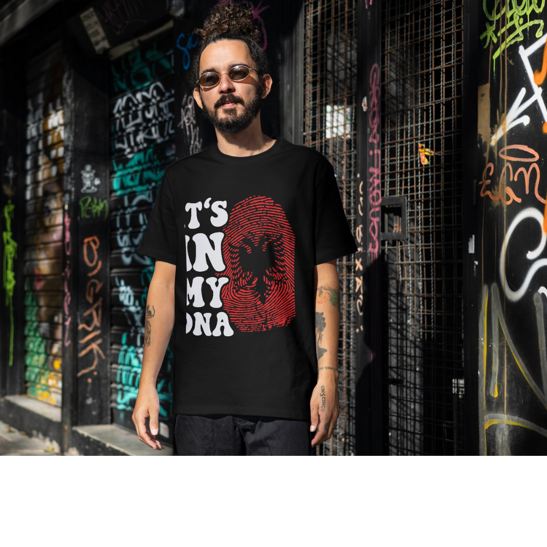 Man wearing a black t-shirt with Albania Flag design and text, standing in front of graffiti-covered walls.